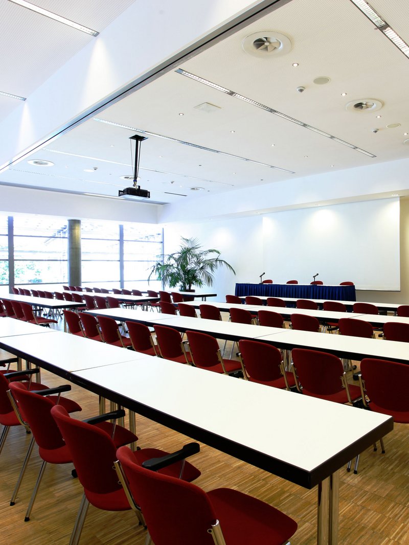 Parlamentarisch bestuhlter Tagungsraum im RuhrCongress Bochum. Der Raum ist von Tageslicht erhellt. Im Hintergrund befindet sich eine große Leinwand, daneben eine Grünpflanze.