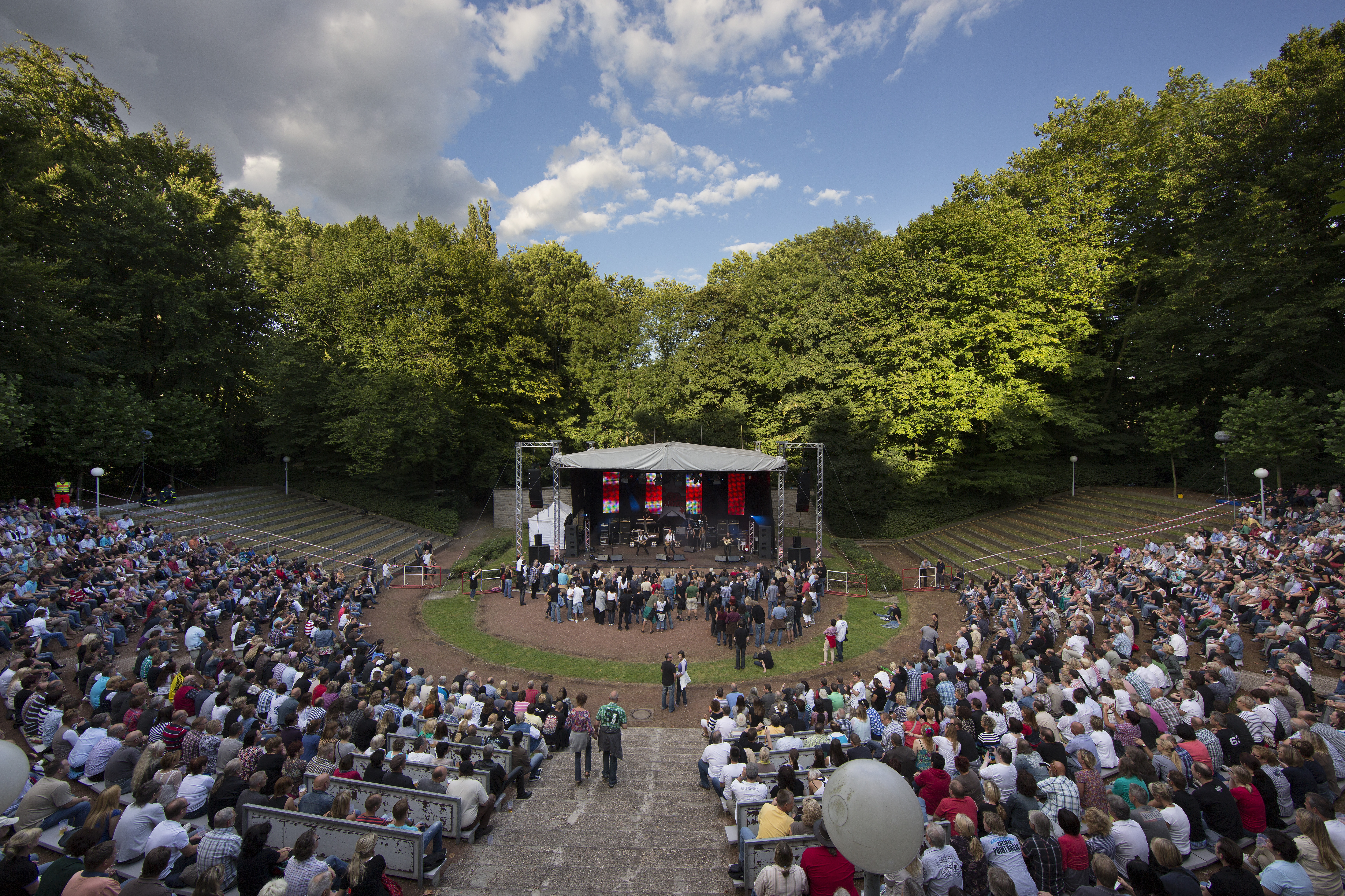 Veranstaltung auf der Freilichtbühne Wattenscheid. Blick von oben, die vollen Ränge hinunter. Einige Menschen Tanzen vor der Bühne. Auf der Bühne sieht man die Band.